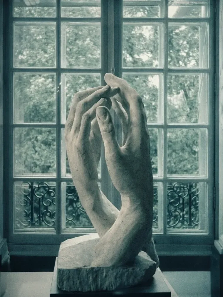 Escultura em pedra de duas mãos que se encontram sem se tocar, diante de uma janela com vista para as árvores